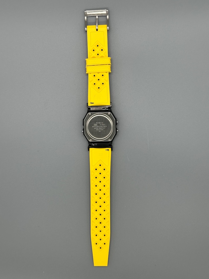 Pode incluir: Um rel&oacute;gio digital preto com uma pulseira amarela brilhante perfurada. O mostrador do rel&oacute;gio &eacute; quadrado e a pulseira &eacute; presa com uma fivela prateada. O rel&oacute;gio &eacute; exibido em um fundo cinza.