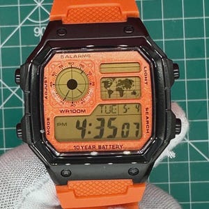 May include: A digital wristwatch with a bright orange resin strap and a black rectangular case. The orange watch face displays a world map, a circular analog-style sub-dial, and the digital time 4:35, along with text reading 5 ALARMS, WR100M, and 10 YEAR BATTERY.