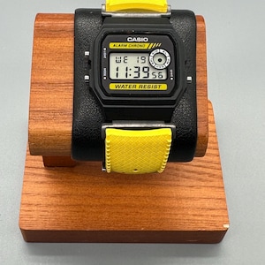 Pode incluir: Um rel&oacute;gio digital Casio preto com uma pulseira amarela &eacute; exibido em um suporte de madeira. O mostrador do rel&oacute;gio mostra a hora 11:39:56. O rel&oacute;gio tem as palavras "CASIO", "ALARM CHRONO" e "WATER RESIST" impressas.