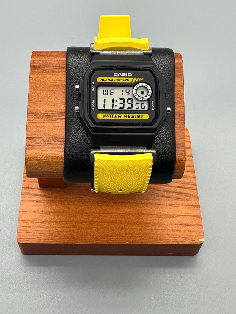 Pode incluir: Um rel&oacute;gio digital Casio preto com uma pulseira amarela &eacute; exibido em um suporte de madeira. O mostrador do rel&oacute;gio mostra a hora 11:39:56. O rel&oacute;gio tem as palavras "CASIO", "ALARM CHRONO" e "WATER RESIST" impressas.