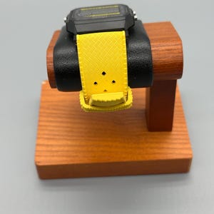 Pode incluir: Um rel&oacute;gio digital preto com uma pulseira amarela &eacute; exibido em um suporte de madeira para rel&oacute;gios. A pulseira do rel&oacute;gio &eacute; de cor amarelo brilhante e tem um padr&atilde;o texturizado. O mostrador do rel&oacute;gio &eacute; preto com um visor amarelo. O suporte &eacute; feito de madeira com acabamento marrom escuro.