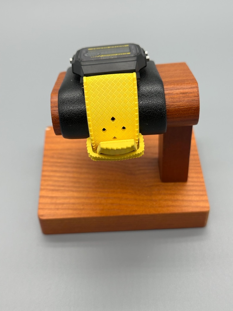 Pode incluir: Um rel&oacute;gio digital preto com uma pulseira amarela &eacute; exibido em um suporte de madeira para rel&oacute;gios. A pulseira do rel&oacute;gio &eacute; de cor amarelo brilhante e tem um padr&atilde;o texturizado. O mostrador do rel&oacute;gio &eacute; preto com um visor amarelo. O suporte &eacute; feito de madeira com acabamento marrom escuro.