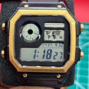 Op de afbeelding: Zwart en goud digitaal horloge met een rechthoekige wijzerplaat. Het horloge geeft de tijd, een wereldkaart en andere functies weer. Het horloge heeft gouden accenten rond de wijzerplaat en op de knoppen.
