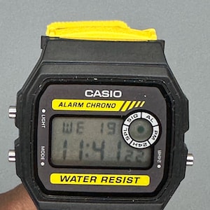Pode incluir: Um rel&oacute;gio digital Casio preto com uma pulseira amarela. O mostrador do rel&oacute;gio exibe a hora e a data, com o texto "CASIO ALARM CHRONO" e "WATER RESIST". O rel&oacute;gio tem formato retangular com cantos arredondados.