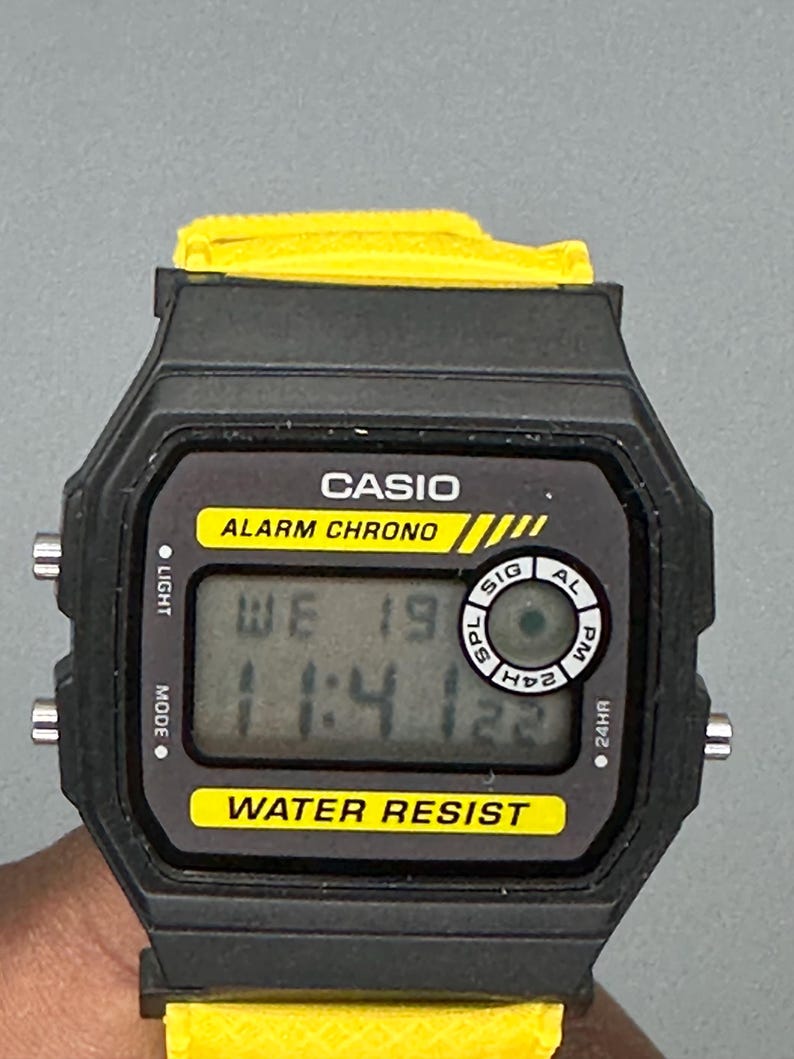 Pode incluir: Um rel&oacute;gio digital Casio preto com uma pulseira amarela. O mostrador do rel&oacute;gio exibe a hora e a data, com o texto "CASIO ALARM CHRONO" e "WATER RESIST". O rel&oacute;gio tem formato retangular com cantos arredondados.