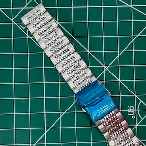 May include: A silver-toned watch band with a mesh-like design. The band is made of small, interconnected metal links. A blue protective film covers the clasp. The band is positioned diagonally against a green grid background.