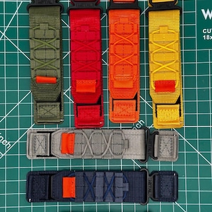 May include: An assortment of tactical straps in various colors, including olive green, red, orange, and yellow. Each strap features a black buckle and orange accent stitching. The straps are arranged on a green cutting mat.