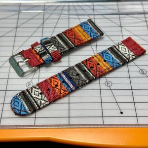 Op de afbeelding: Twee kleurrijke horlogebandjes met een geweven patroon in rood, oranje, blauw en zwart. De bandjes hebben een geometrisch ontwerp en een zilveren gesp. Ze liggen op een witte snijmat.