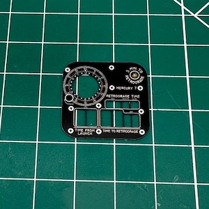 Puede incluir: Un componente negro de forma cuadrada con intrincadas marcas blancas, que incluyen un indicador circular, ventanas rectangulares y texto que dice "5 MIN TO RETROGRADE" y "TIME FROM LAUNCH". El componente está sobre una cuadrícula verde.