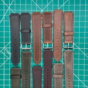 May include: An assortment of leather watch straps in various colors, including black, brown, and gray. Each strap features a buckle and stitching detail. The straps are laid out on a green cutting mat.