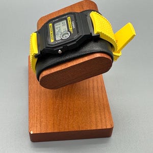 Pode incluir: Um rel&oacute;gio digital Casio preto com uma pulseira amarela &eacute; exibido em um suporte de rel&oacute;gio de madeira. O mostrador do rel&oacute;gio mostra a hora. O suporte &eacute; feito de madeira tingida e tem uma almofada de couro preta.