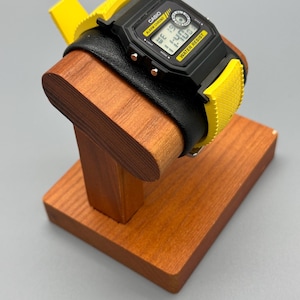 Pode incluir: Um rel&oacute;gio digital preto com detalhes amarelos e uma pulseira de couro preta &eacute; exibido em um suporte de madeira para rel&oacute;gios. O mostrador do rel&oacute;gio mostra a hora. O suporte &eacute; feito de madeira tingida.