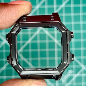 May include: A dark gray watch case with a square face and rounded corners. The case has a clear window and several small, circular buttons on the sides. The watch case is held by a person's fingers.