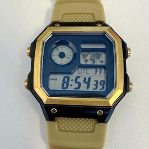 May include: A digital wristwatch with a gold-colored bezel and a tan band. The watch face is dark blue with a digital display showing the time as 8:54:39 PM, a world map, and other indicators. The watch has gold-colored buttons.