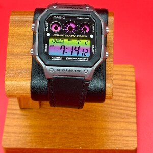 May include: A digital Casio watch with a black leather strap. The watch face shows the time, day, and date. The watch has a silver and black case with buttons for adjusting the mode, light, and start functions. The watch is displayed on a wooden stand.