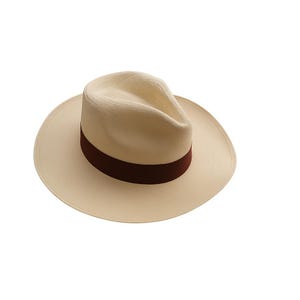 Op de afbeelding: Een crèmekleurige fedora hoed met een brede rand en een donkerbruine band. De hoed heeft een klassiek ontwerp en is geschikt voor diverse gelegenheden. De hoed is gemaakt van een zacht materiaal.