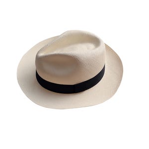 Könnte beinhalten: Ein cremefarbener Panamahut mit schwarzem Band. Der Hut hat eine klassische Fedora-Form mit breiter Krempe. Der Hut ist aus gewebtem Stroh gefertigt.