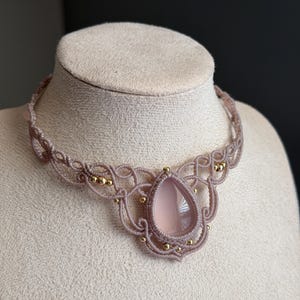 May include: A macrame choker necklace featuring a central teardrop-shaped light pink stone. The necklace is crafted from light brown cord in an intricate knotted pattern, accented with small, round, polished gold-colored metal beads.