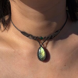 May include: A macrame choker necklace featuring a teardrop-shaped labradorite gemstone pendant with iridescent green and yellow flashes. The dark brown cord is intricately knotted and includes a small green bead accent at the center.