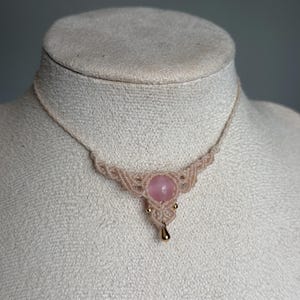 May include: A macrame necklace featuring a central round pink stone, accented by two small gold-toned beads and a single gold-toned teardrop charm. The cord is crafted from light beige thread, woven into a decorative pattern around the stone.