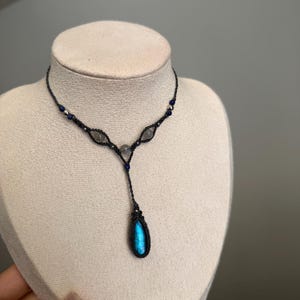 May include: A macrame necklace featuring a dark cord, small blue and silver beads, and three light-colored stones. A central teardrop-shaped pendant with a vibrant blue iridescent stone hangs from the bottom of the Y-shaped design.