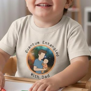 May include: A smiling toddler wearing a beige t-shirt with the text "Quantum Entangled With Dad" and a graphic of a father and child. The child is sitting at a wooden table with crayons and paper.
