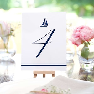 May include: A white table number card with a navy blue sailboat illustration and the number 4. The card is displayed on a small wooden easel. A navy blue stripe is at the bottom of the card.