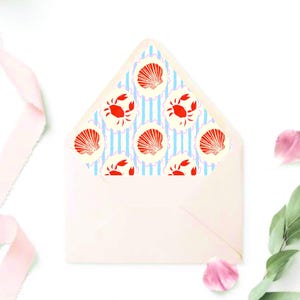 May include: A white envelope with a patterned interior featuring red crabs and seashells on a blue and white striped background. The envelope is decorated with pink ribbon, flower petals, and green leaves.