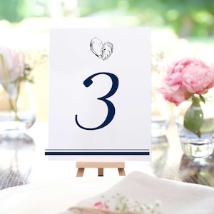 May include: White table number card with a navy blue border and a navy blue number "3". The card has a small illustration of two oyster shells in the shape of a heart. The card is on a wooden easel.