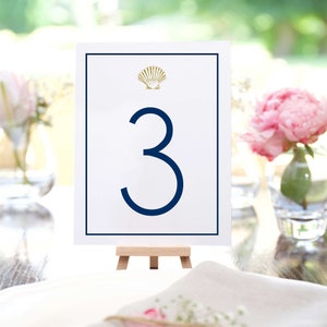 May include: White table number card with a navy blue border and a gold seashell design. The number "3" is printed in navy blue on the card. The card is displayed on a small wooden easel.