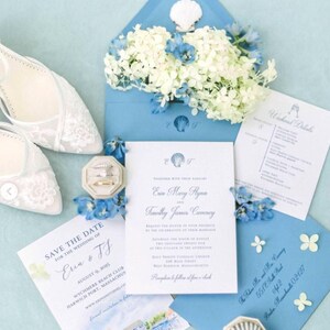May include: A wedding invitation suite featuring a blue envelope with a seashell, white flowers, and a wedding invitation. The suite includes a "Save the Date" card, a details card, and a pair of white lace wedding shoes. The invitation text includes the names Erin and Timothy.