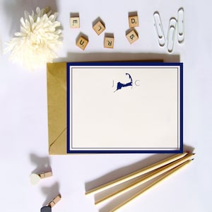 May include: A white card with a navy blue border and a navy blue outline of the Cape Cod peninsula with the letters J and C on either side. The card is on a white surface with a brown envelope, wooden pencils, and white and black paper clips.