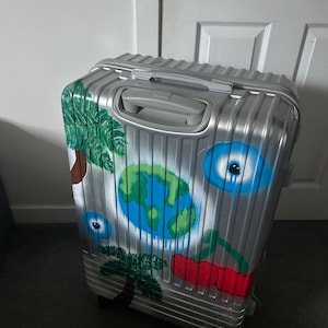 May include: A silver travel suitcase with a painted design. The artwork features a blue and green globe, palm trees, cherries, and blue eye designs. The suitcase has a retractable handle and wheels.