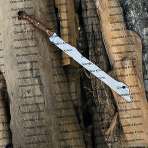 May include: A long, silver blade with a brown and tan handle. The blade is straight and wide, with a slight curve at the tip. The handle is intricately designed. The text "Grandeur Knives" is repeated in the background.