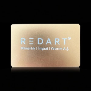 Puede incluir: Una tarjeta de visita rectangular de color dorado con la palabra "REDART" en letras azul claro. Debajo del nombre hay palabras adicionales en azul claro. La tarjeta tiene una textura de metal cepillado y está sobre un fondo negro.