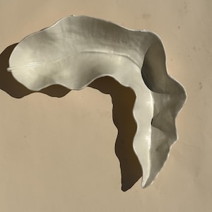 May include: A cream-colored ceramic dish shaped like a curved, elongated leaf. The piece features a smooth, glossy finish with a central vein detail and undulating, organic edges that create a deep, concave form for holding small items.