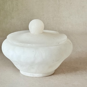 May include: A white stone bowl with a matching lid featuring a spherical handle. The bowl has a rounded, wide body with a textured, marbled surface and a smooth, circular base. The lid sits securely on top, completing the vessel's form.