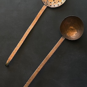 May include: Two copper kitchen utensils with long, slender handles featuring a repeating etched chevron pattern. One utensil is a round, perforated skimmer, and the other is a solid, deep, rounded ladle with a hammered texture finish.