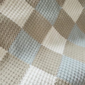 May include: A handmade crochet blanket with a diamond pattern. The blanket features squares of white, light blue, and various shades of brown. The textured surface of the blanket is visible in this close-up shot.