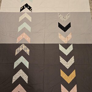 May include: A rectangular quilt featuring a color-blocked design in white, light gray, and dark gray. Two vertical columns of chevron-shaped fabric patches in various patterns and colors, including mint, black, gold, and pink, run down the quilt.