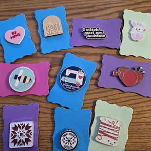 May include: A collection of colorful enamel pins on decorative foam cutouts. The pins feature various designs, including a heart with "Be Mine", a rainbow, a bee, a sewing machine, a pumpkin, a bunny, a quilt block, a needlepoint hoop, and a spool of thread.