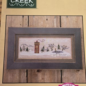 May include: A cross-stitch pattern titled "Long Winter" by Bent Creek, featuring a winter landscape with a red building, trees, and snow. The design is framed in a dark gray frame. The book number is BC1076.