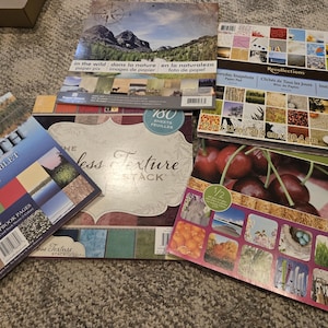 May include: A collection of four square scrapbook paper pads featuring various designs including mountain landscapes, cherry patterns, and textured backgrounds. The pads display text such as North Scrapbook Tablet, The Timeless Texture Stack, and Recollections.