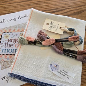May include: A cross-stitch kit with a white Aida cloth, embroidery floss in various colors, and a pattern booklet. The booklet reads "sweet wing studio" and "enjoy the moment". The floss colors include shades of pink, brown, and blue.