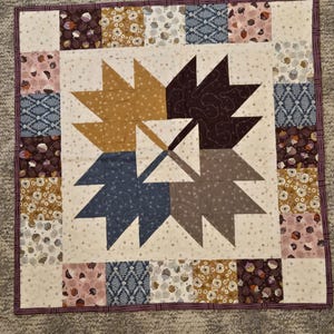 May include: A square patchwork quilt featuring a central bear paw block design in mustard, dark brown, blue, and gray. The border consists of alternating squares with floral, acorn, and fern patterns on cream backgrounds, finished with a dark purple binding.