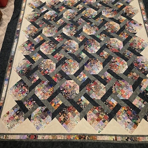 May include: A rectangular quilt featuring a repeating pattern of octagonal blocks composed of various multi-colored floral and patterned fabric squares. The octagons are interconnected by dark grey fabric strips, framed by a cream border.