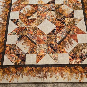 May include: A square quilt featuring a central star pattern composed of fabric squares with autumn leaf prints in shades of orange, brown, and gold, interspersed with cream-colored squares. A wide border displays a detailed fall foliage motif.
