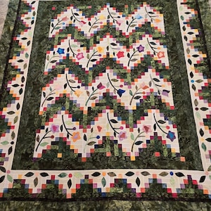 May include: A rectangular quilt featuring a central pattern of zigzagging green and white fabric strips interspersed with colorful floral appliqués. The border consists of a dark green leafy print framed by a white band with small flower motifs.
