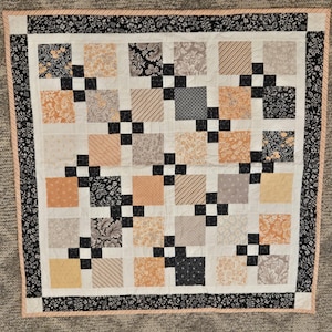 May include: A square quilt featuring a patchwork design of alternating blocks in shades of peach, cream, gray, and black. The pattern includes floral, striped, and geometric prints, separated by small black squares, with a black floral border.