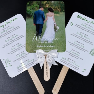 May include: Wedding fan with a photo of a bride and groom, and wedding details. The fan has a white ribbon and wooden handle. The text on the fan says "Welcome to our wedding" and the names Sophia & Michael.